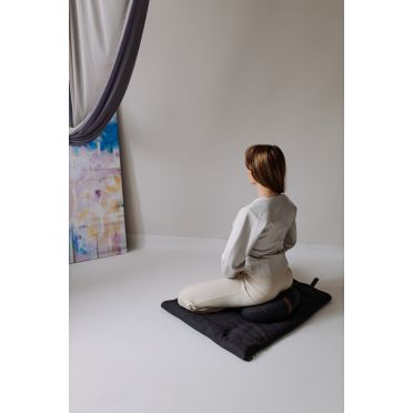 Kobieta siedzi w pozycji medytacyjnej na ciemnoszarej poduszce do medytacji, jogi i relaksacji w kształcie fasolki, ułożonej na dopasowanej macie w minimalistycznym wnętrzu., w tle namalowany obraz.